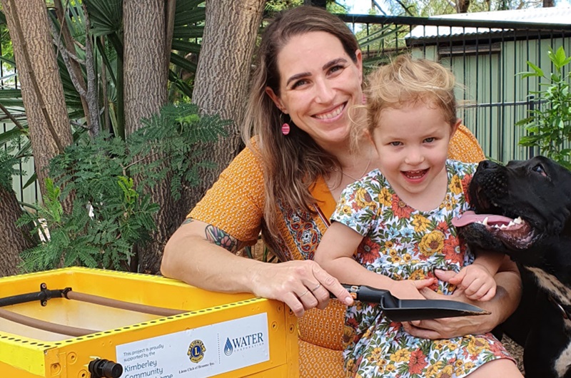 Image of woman with child on lap and dog next to them. They are crouched beside a fridge that has been upcycled into a raised garden bed and hand-painted.