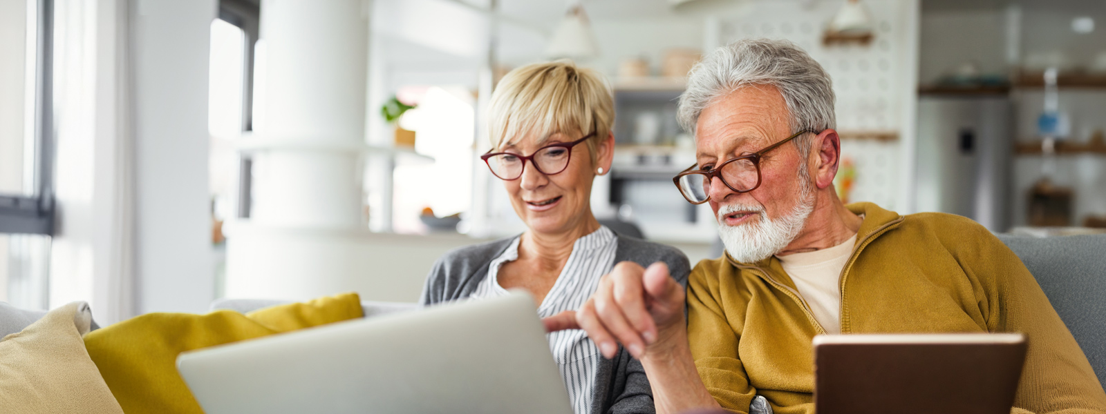 An elderly married couple sitting on their couch and looking something up on their laptops