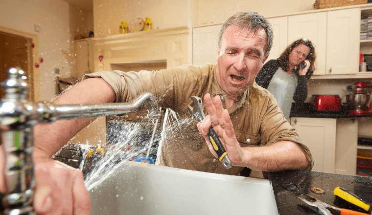 Man trying to fix water bursting out of the kitchen tap. Wife in the background calling a plumber