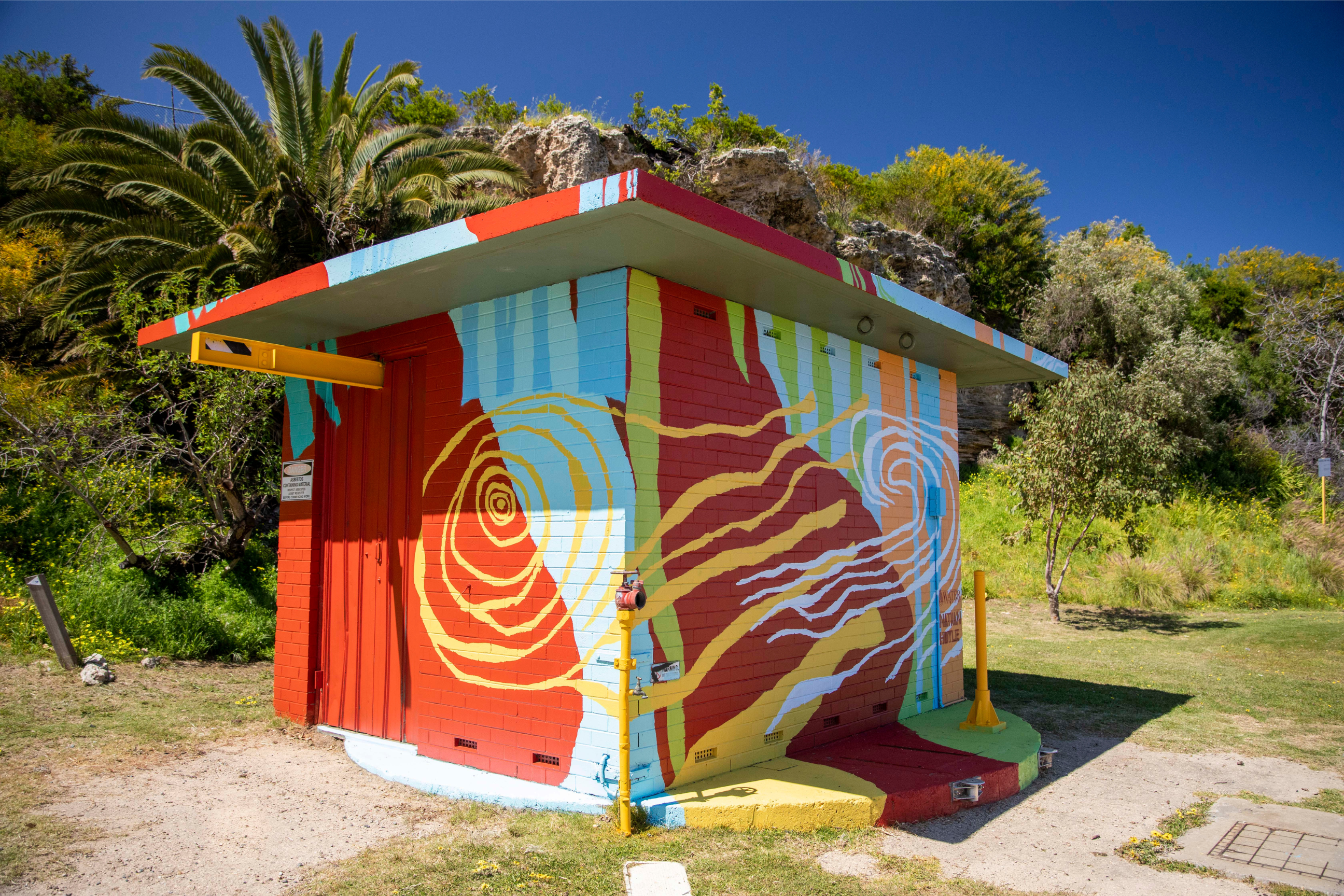 Small utility building covered in a vibrant mural of bold red, blue, yellow, and green shapes with flowing organic patterns, set against coastal vegetation and rocky outcrops.