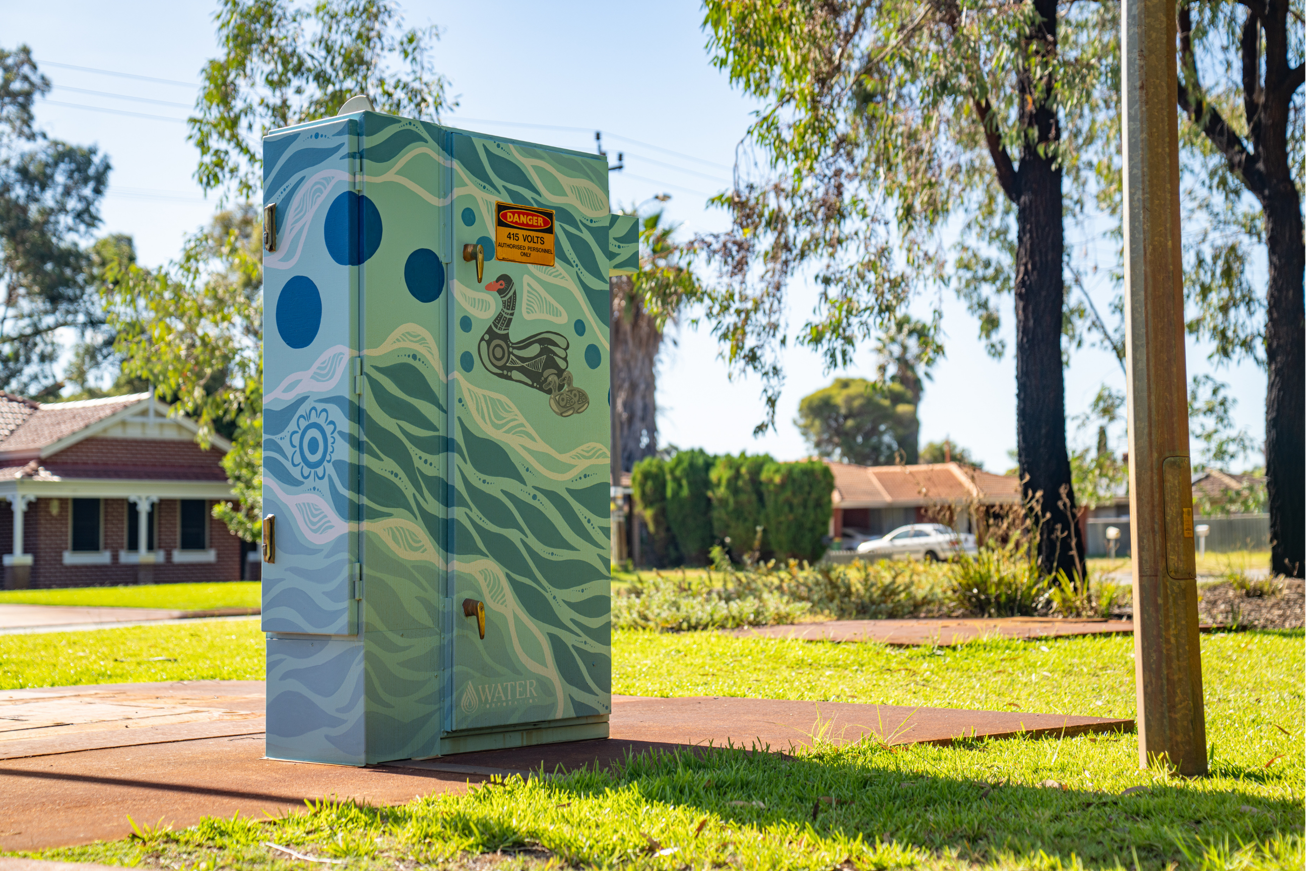 Utility box painted with flowing blue and green patterns, circular motifs, and a stylised bird design, situated in a leafy suburban park.