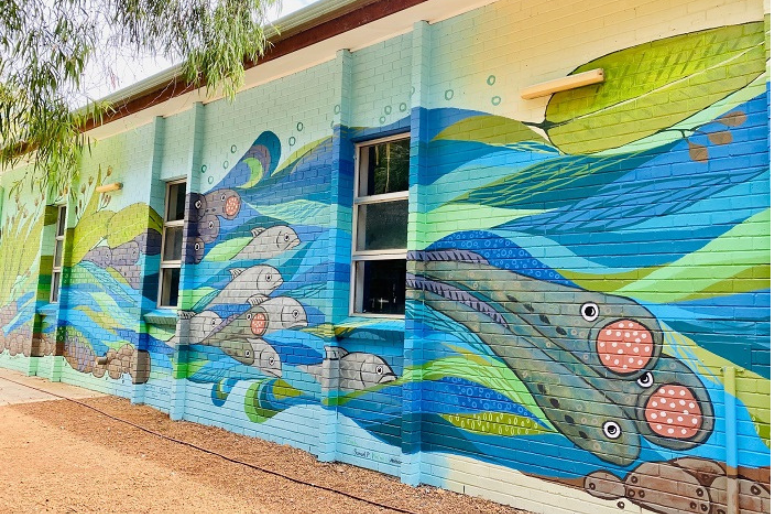 Building wall covered in a vibrant blue and green mural featuring native fish, flowing water patterns, and aquatic plants inspired by a freshwater ecosystem.