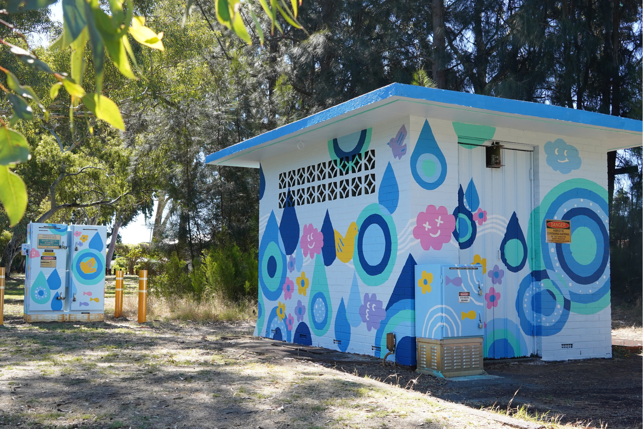 Small utility building painted with cheerful raindrop, flower, bird, and rainbow motifs in shades of blue, teal, pink, yellow, and purple, surrounded by trees.