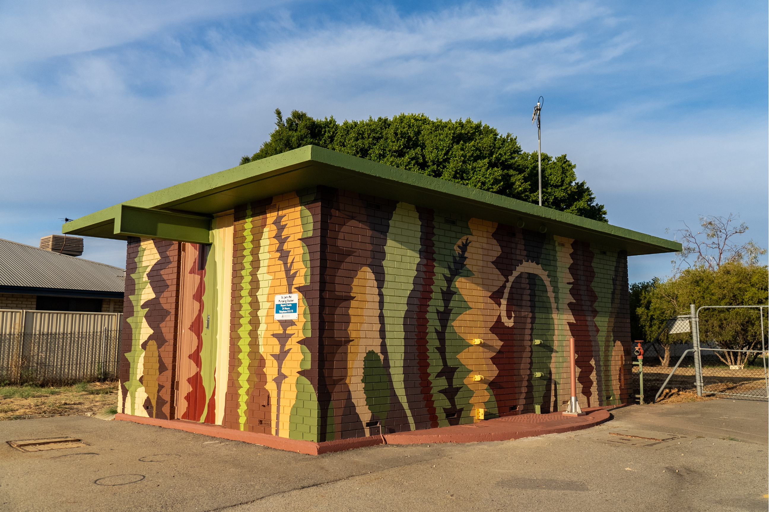 Utility building painted with an abstract mural of layered organic shapes in earthy green, brown, tan, and ochre tones, inspired by the natural landscape.