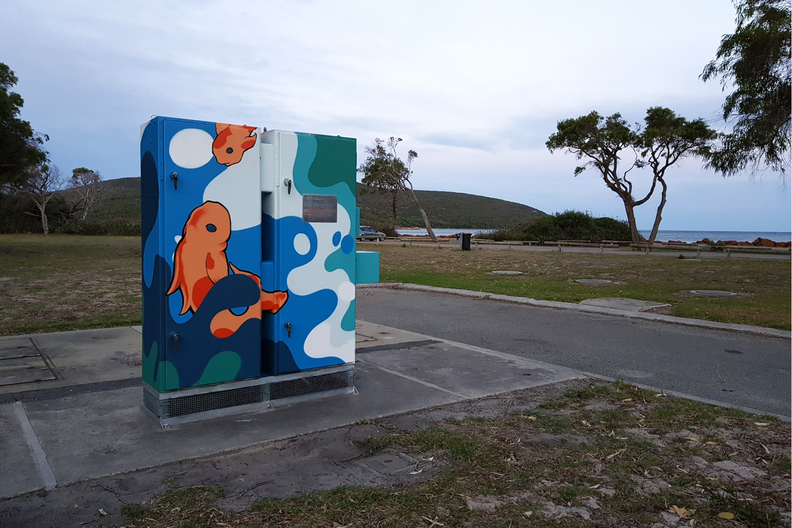 Utility box decorated with a stylised orange fish swimming among flowing blue, teal, and white shapes, set against a coastal landscape and ocean backdrop.