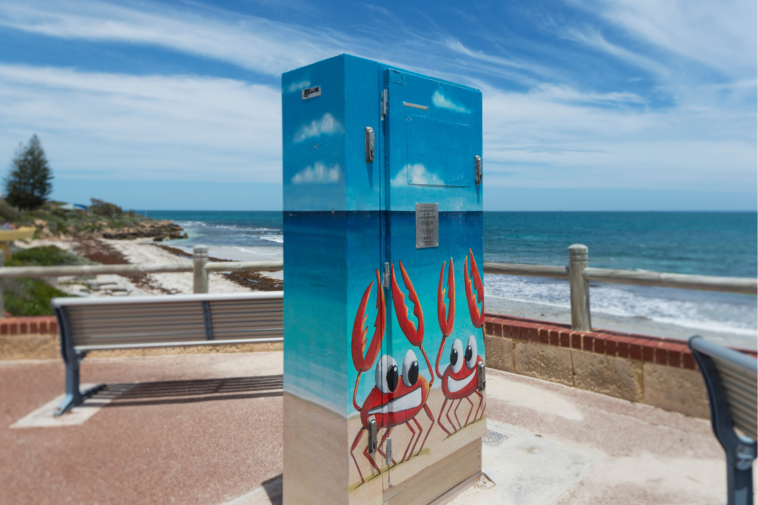 Utility box painted with playful red crabs against a bright blue sky and beach scene, positioned along an oceanfront promenade overlooking the coast.
