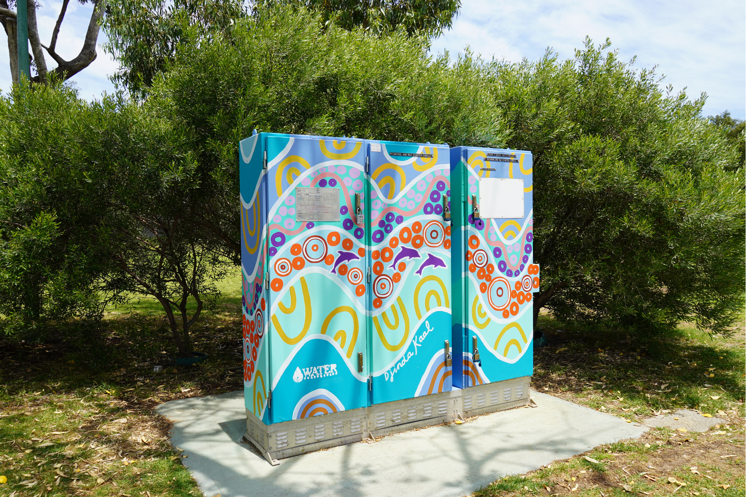 Utility box decorated with colourful wave patterns, circular designs, and dolphin motifs in turquoise, purple, orange, and yellow, nestled among native trees and parkland.