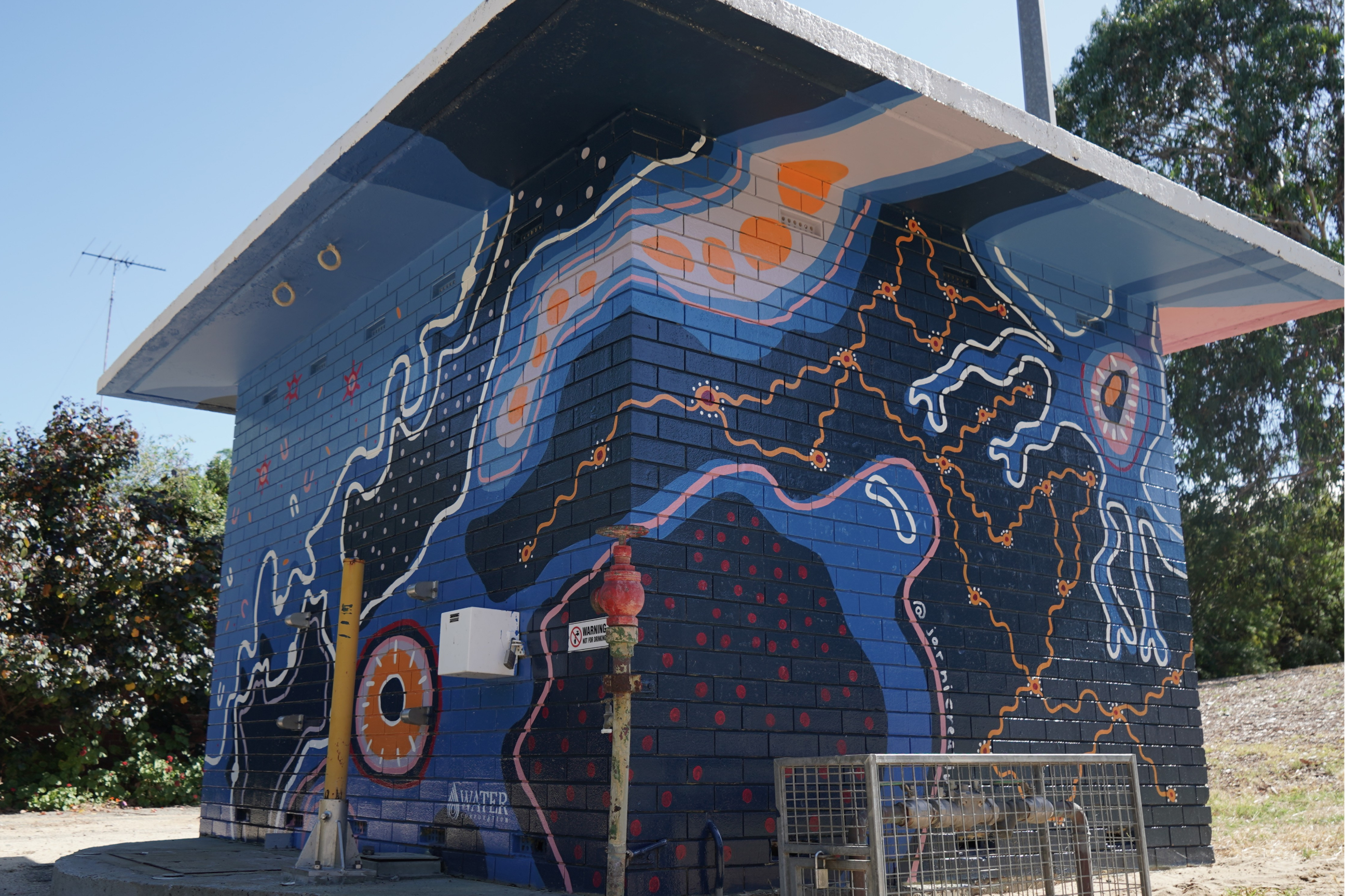 Utility building covered in a striking abstract mural of flowing lines, circular motifs, and organic patterns in deep blue, orange, pink, and white tones.
