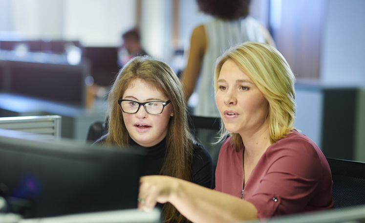 A young school leaver gets training on the computer in a busy office.