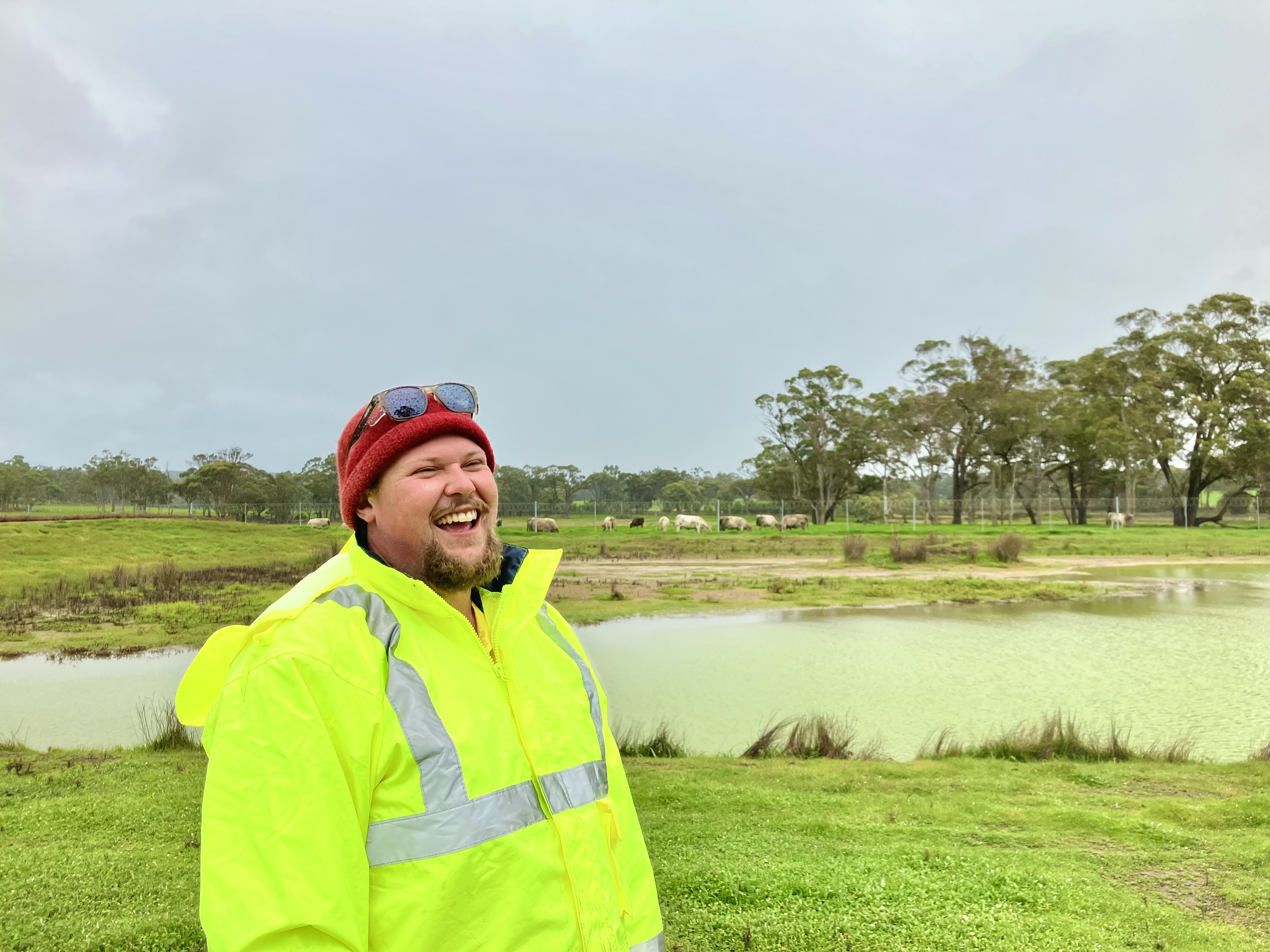 Todd, wearing a yellow high-visibility jacket and red beanie, smiles while standing near a pond in a grassy field with cows grazing in the background under an overcast sky.