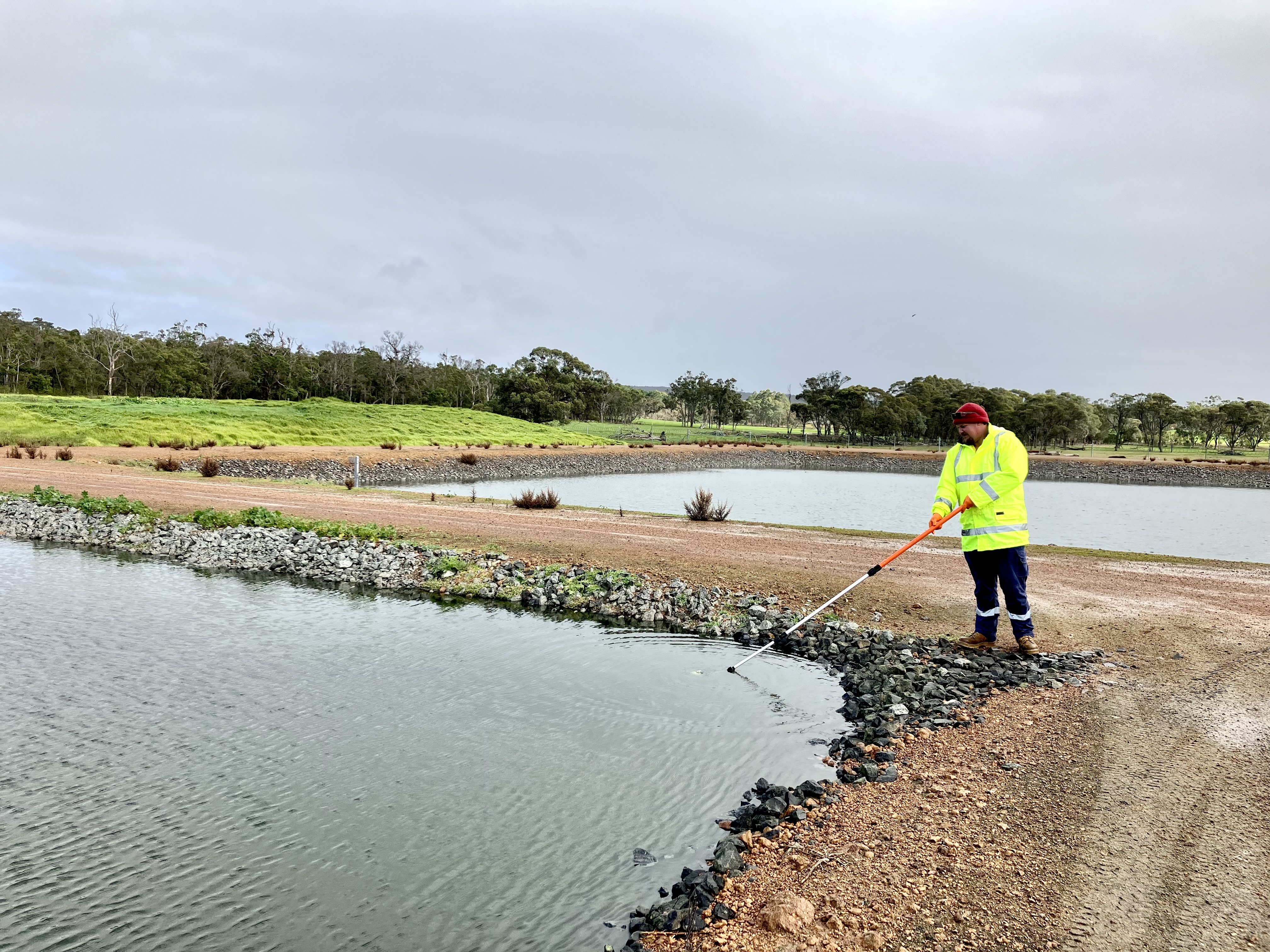 Todd, dressed in a yellow high-visibility jacket and red beanie, uses a long-handled tool to inspect or clean the edge of a pond surrounded by grass, rocks, and trees.