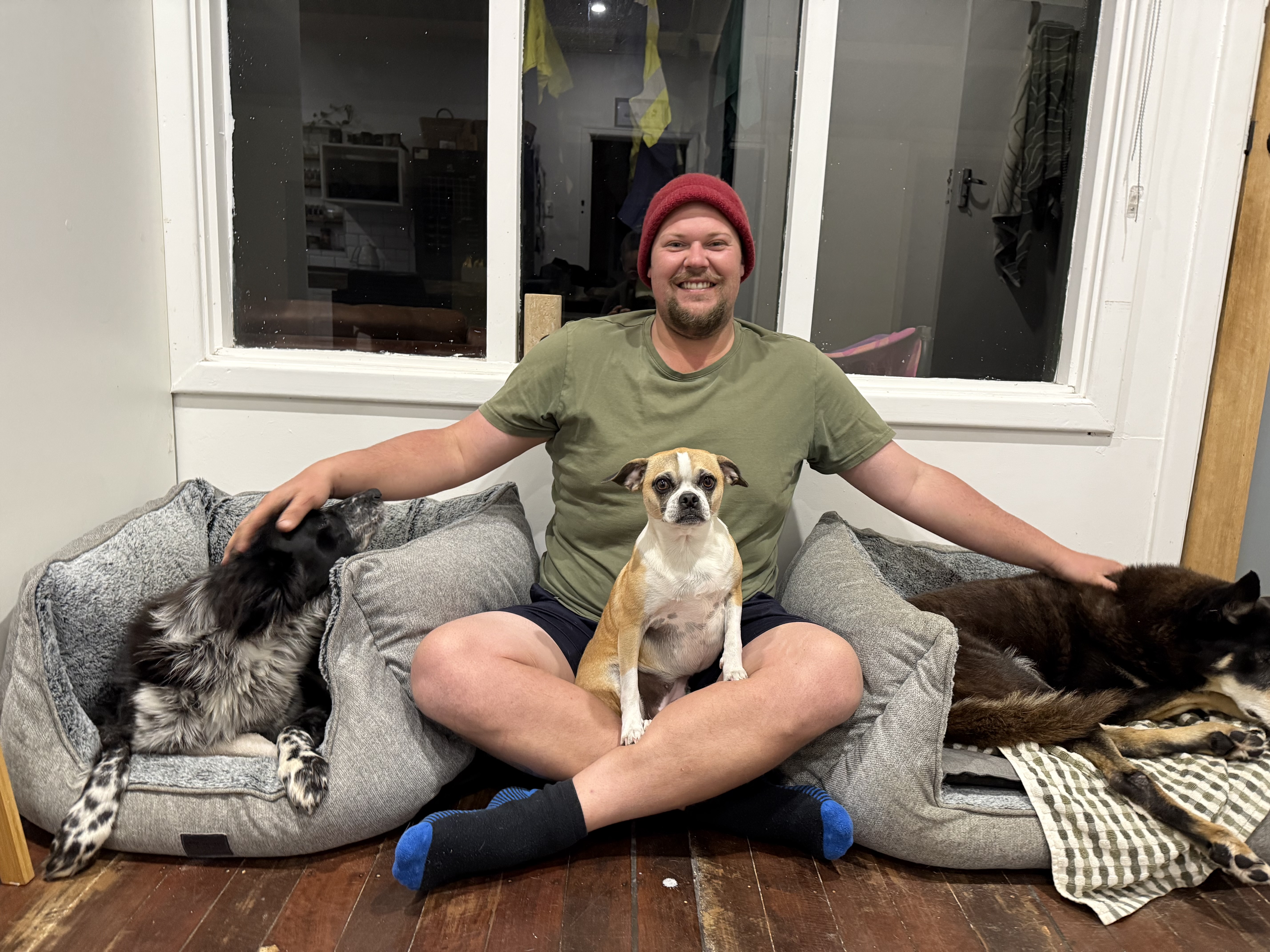 Todd sits indoors on a wooden floor, smiling and petting three dogs—Sam, Frankie, and Pip—two lying in dog beds and one sitting upright between his legs.