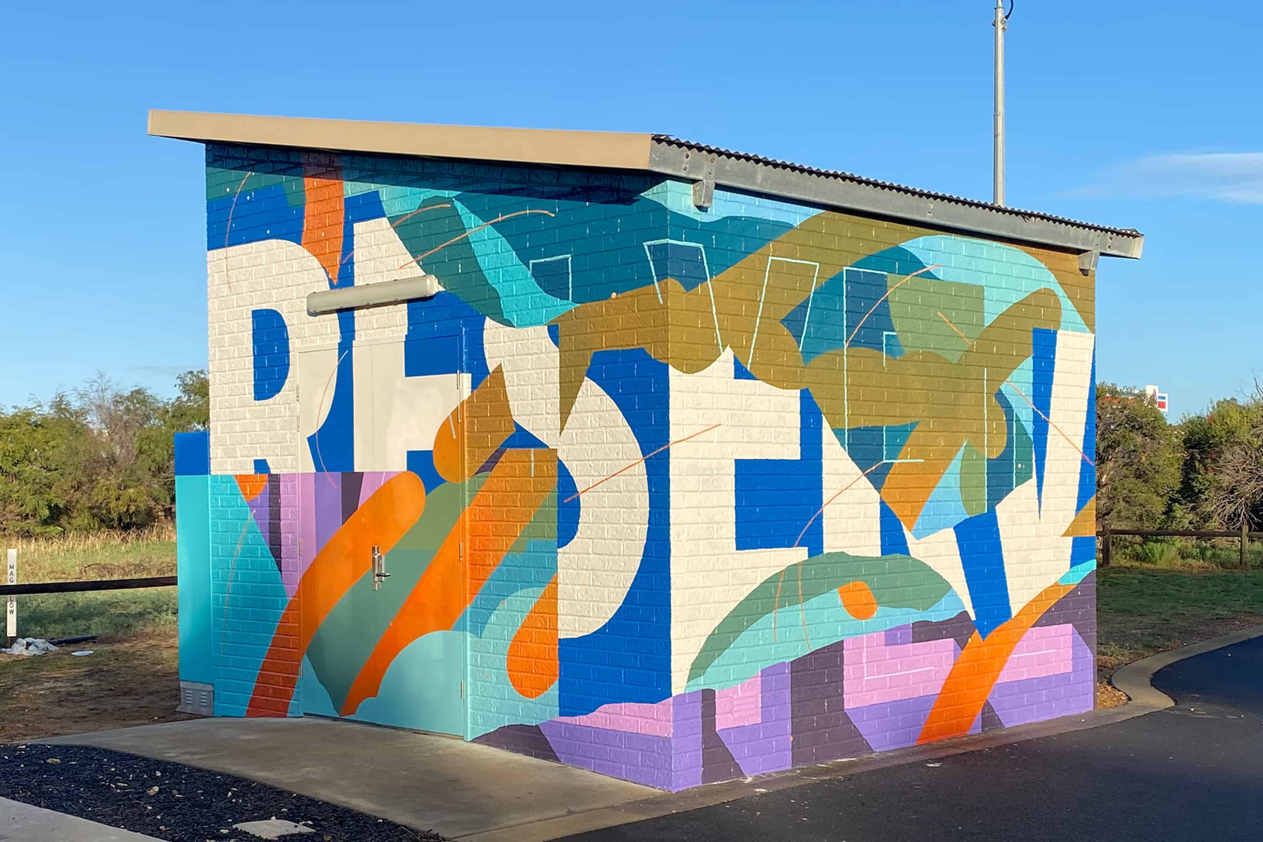 Small utility building transformed with a bold abstract mural of overlapping geometric shapes in blue, orange, teal, purple, and cream tones.