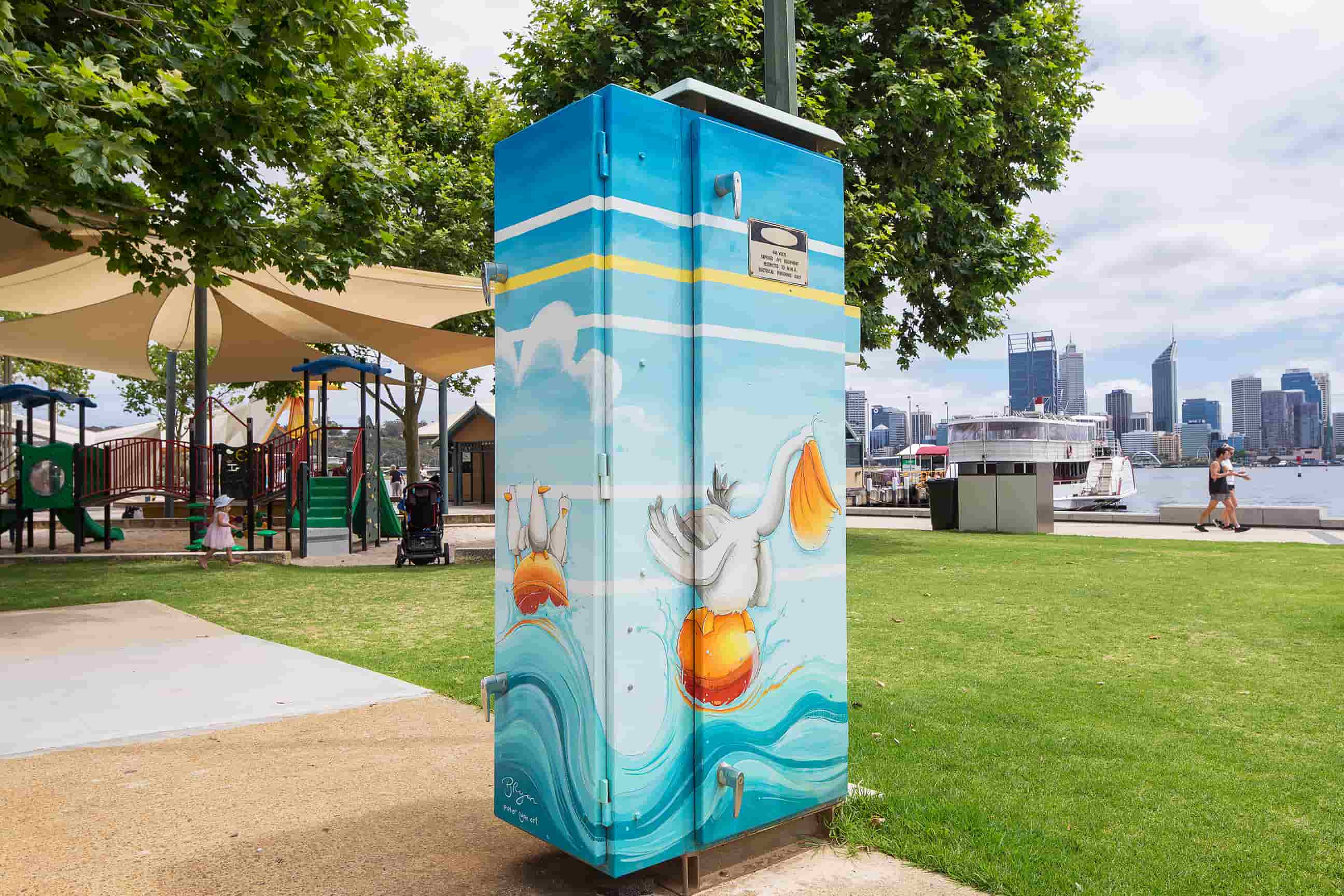 Utility box decorated with playful pelican illustrations floating on turquoise water, set in a waterfront park with a city skyline in the background.