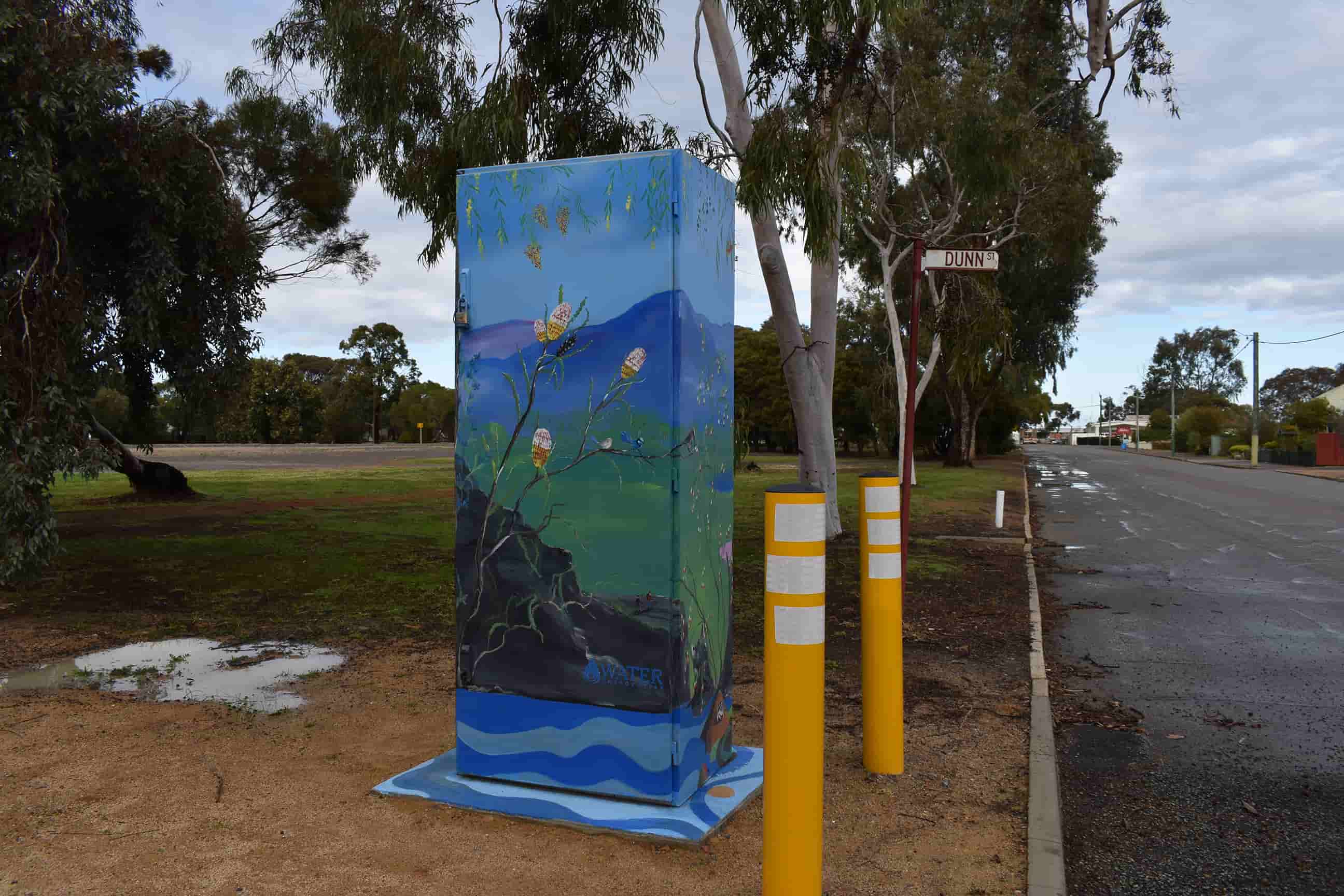 Utility box painted with a serene native landscape mural featuring flowering banksia branches, rolling blue hills, and local flora beside a tree-lined roadway.
