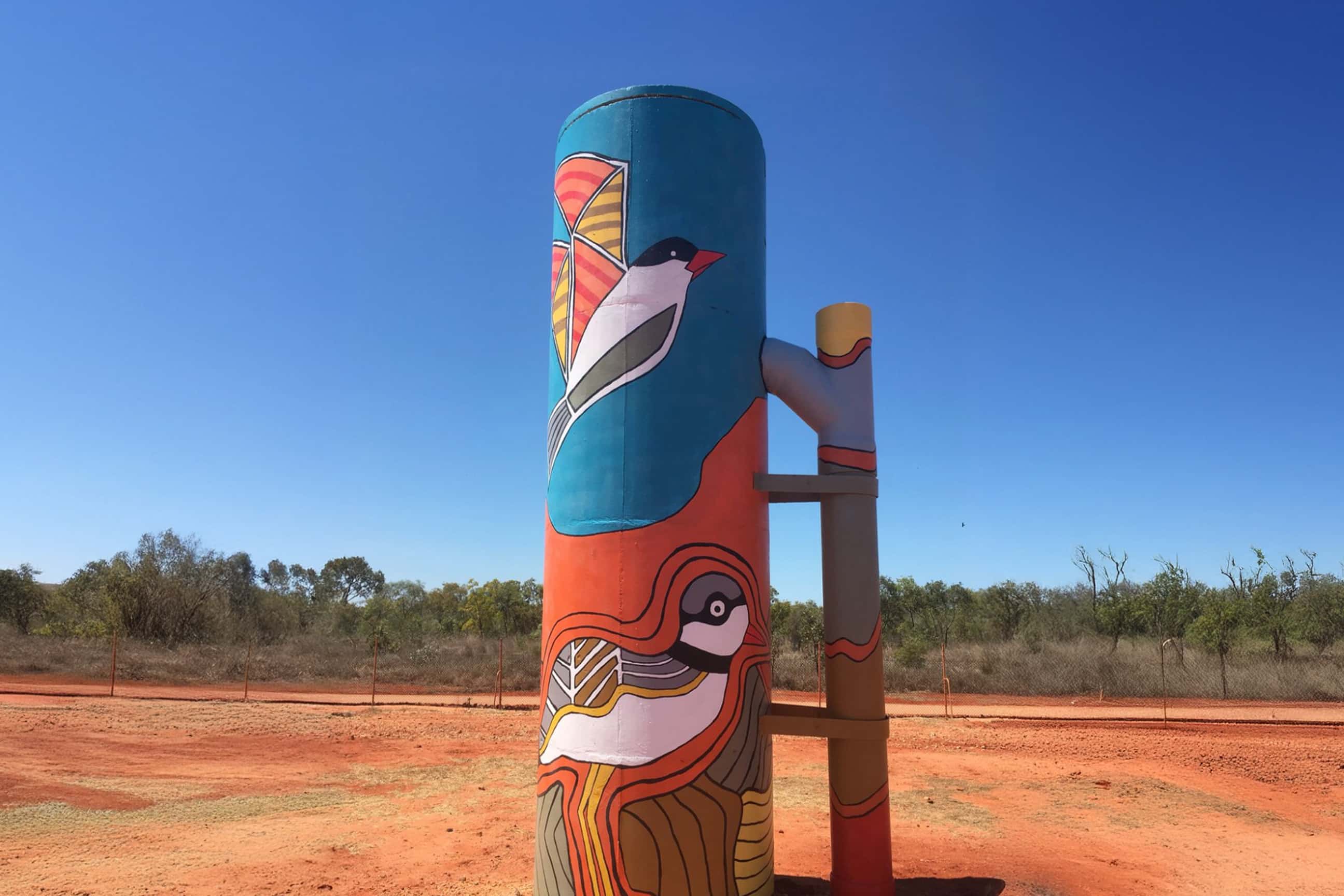 Brightly painted water tank in a red-earth landscape, decorated with stylised bird artwork and vibrant blue, orange, and yellow patterns beneath a clear sky.