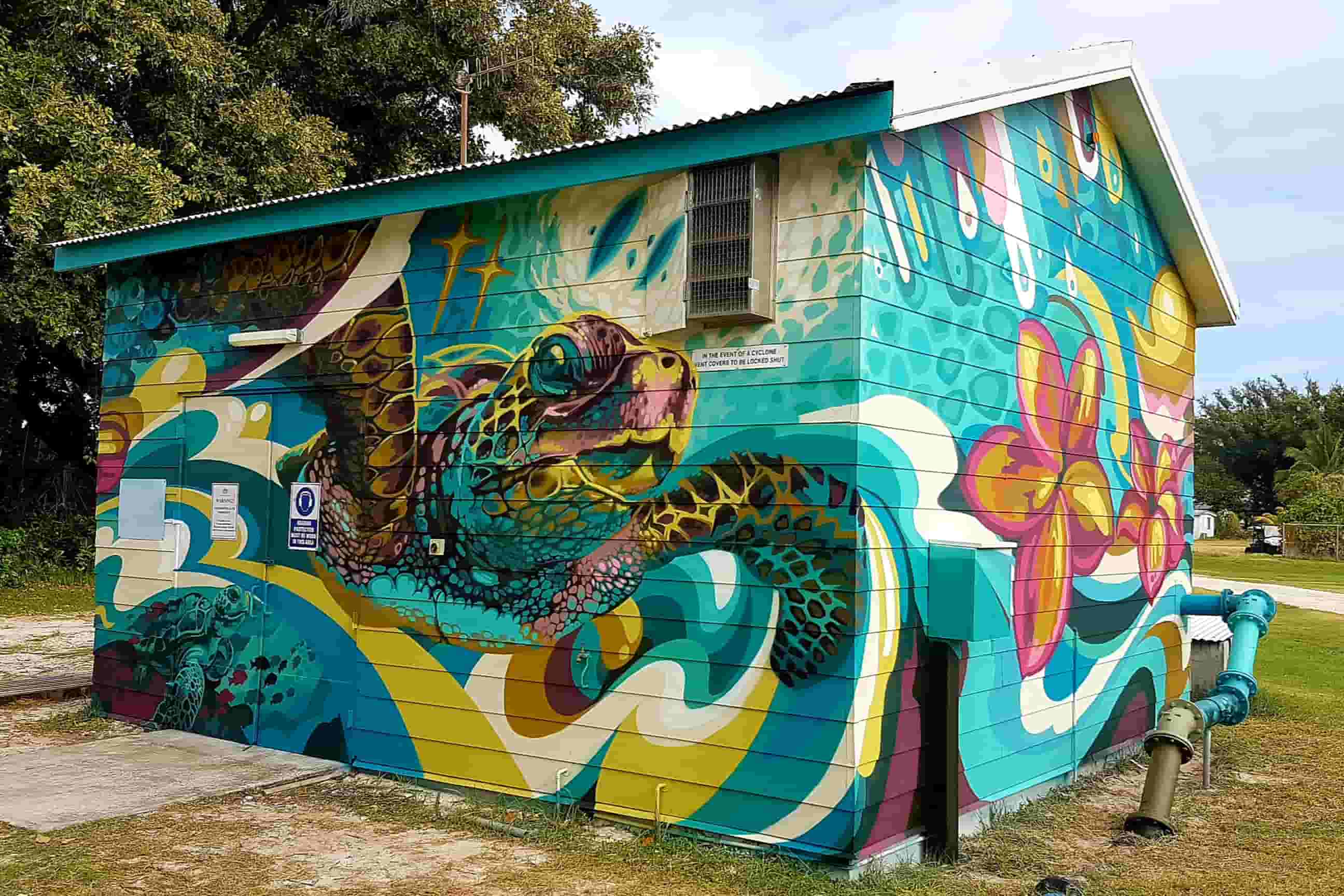 Utility building covered in a vivid mural of a sea turtle swimming through swirling turquoise, yellow, pink, and white ocean-inspired designs.