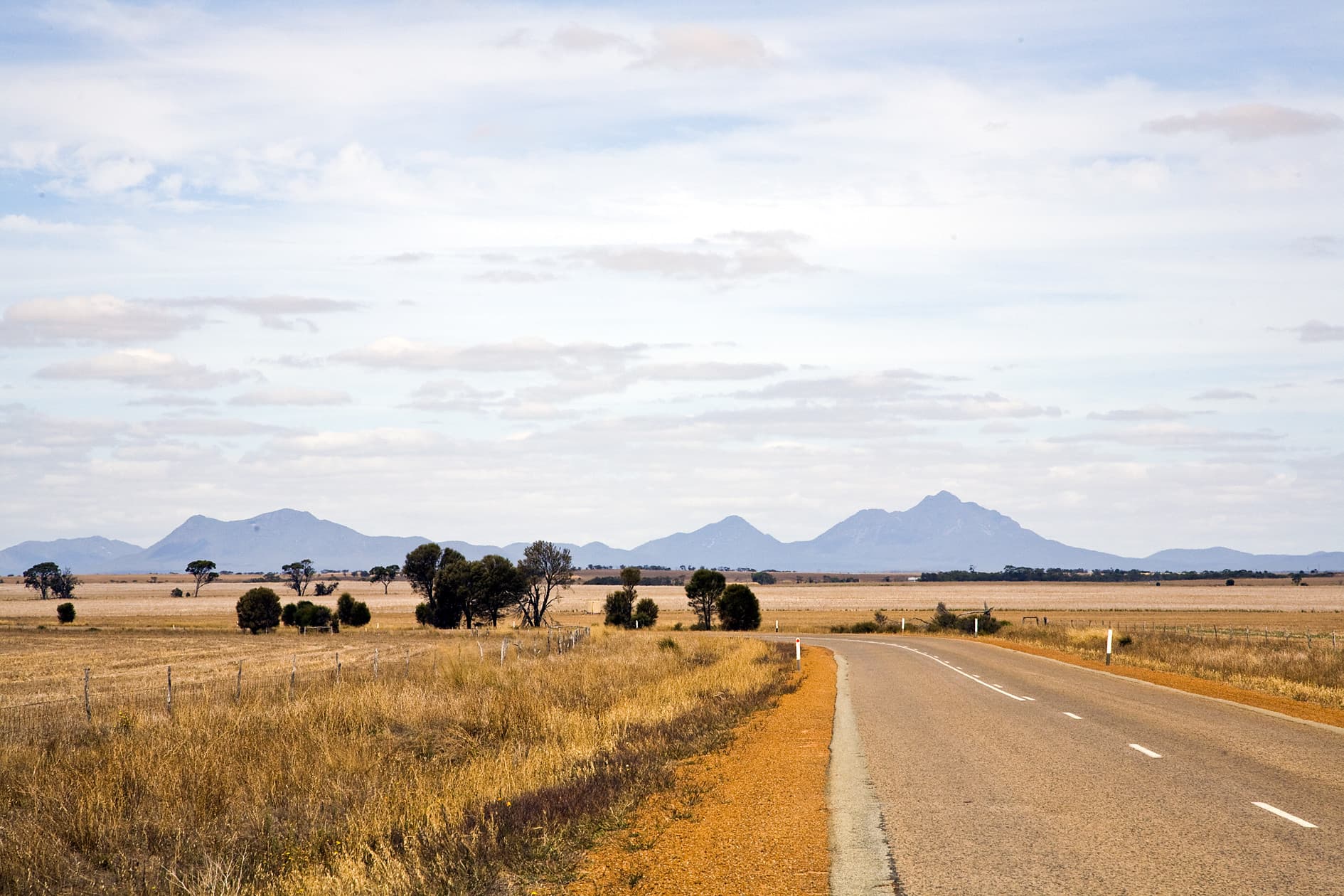 Rural road winding through dry farmland, with golden paddocks, scattered trees and mountain ranges visible in the distance under a partly cloudy sky.