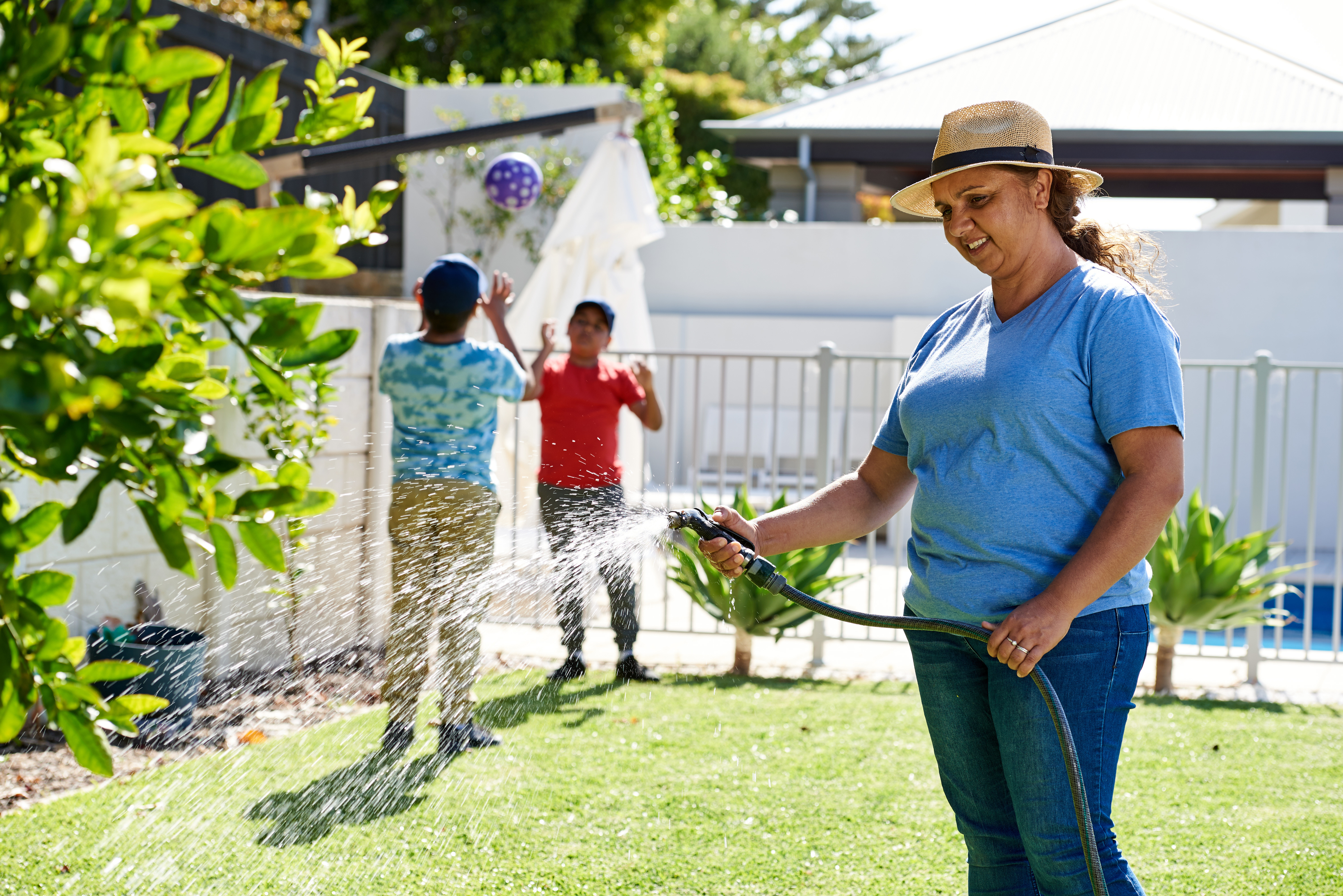 Person watering a backyard garden with a hose while children play on a lawn in the background.